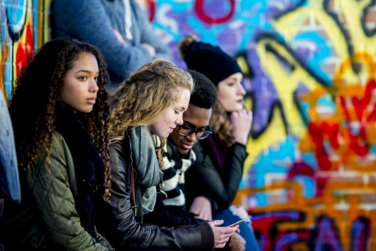 A group of teenagers are sitting in front of a wall covered in graffiti. They are wearing stylish clothes. A boy and girl are looking at a smartphone screen together.