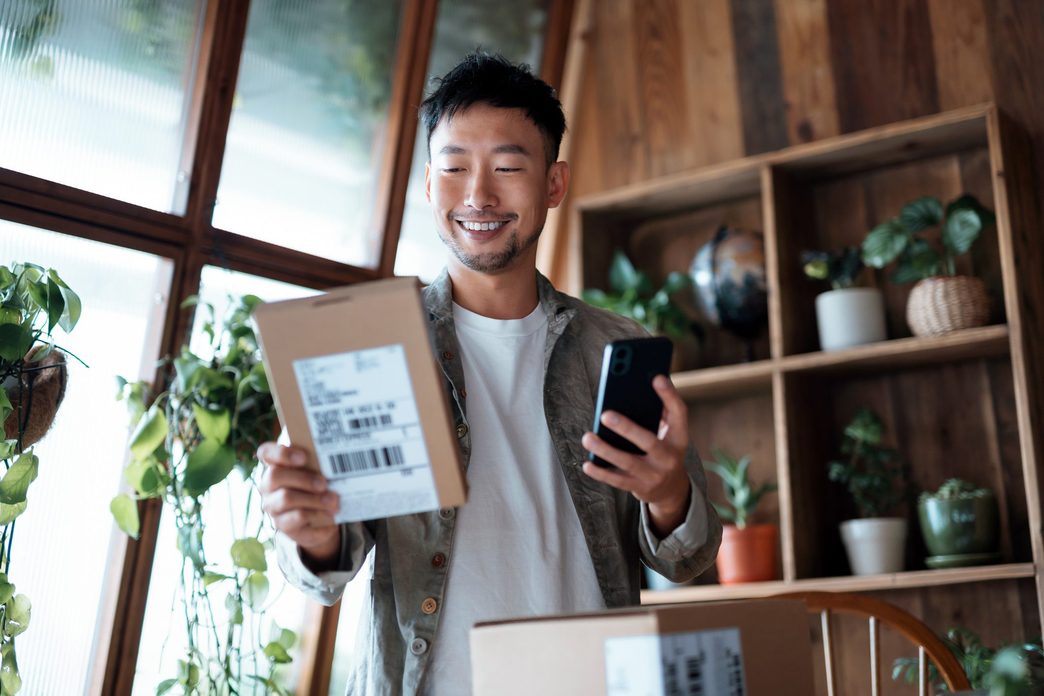 Smiling young Asian man checking his smartphone as he holds delivered package from online purchases at home.