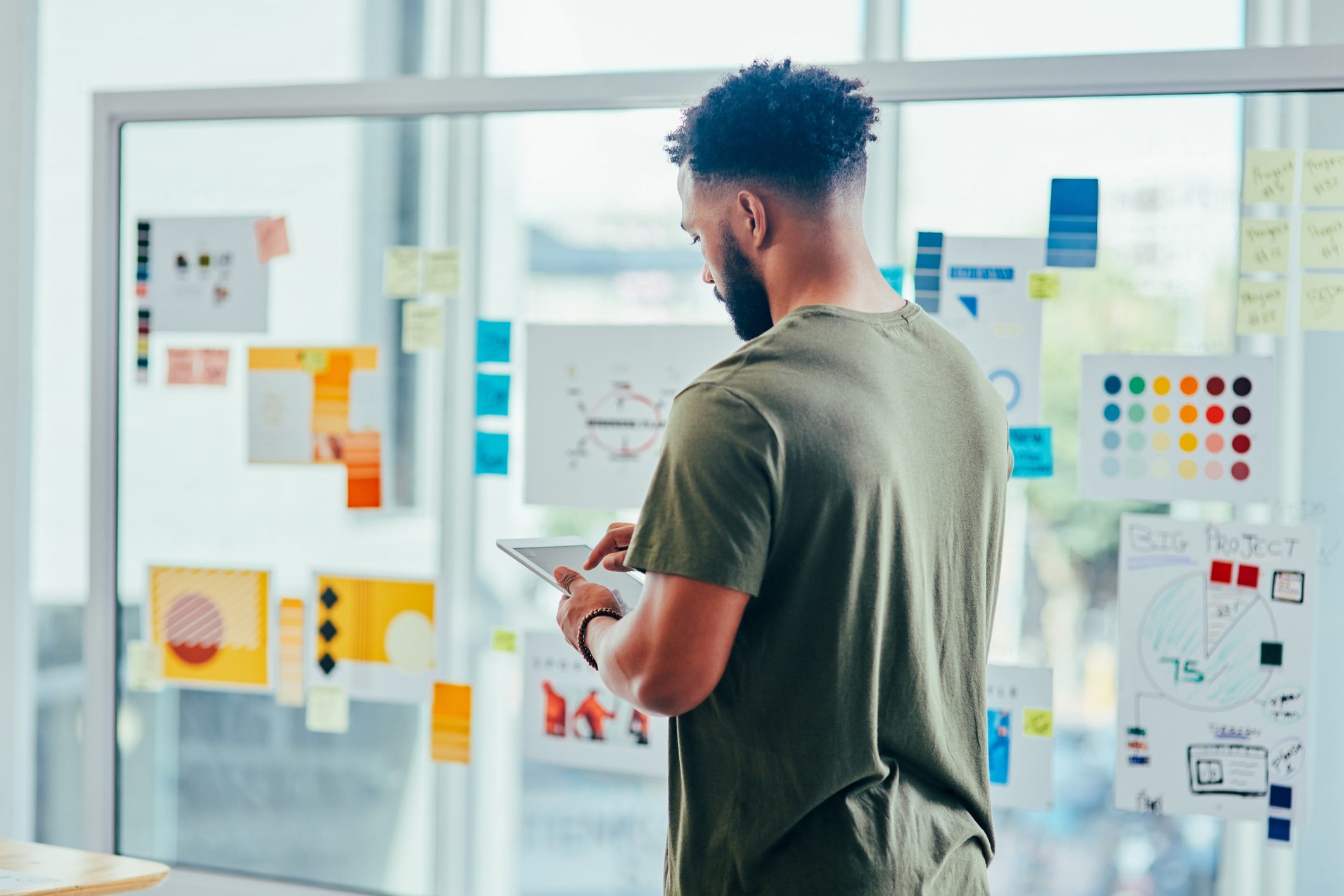 Monitoring market trends, Rearview shot of a young designer using a digital tablet while brainstorming with notes on a glass wall in an office.