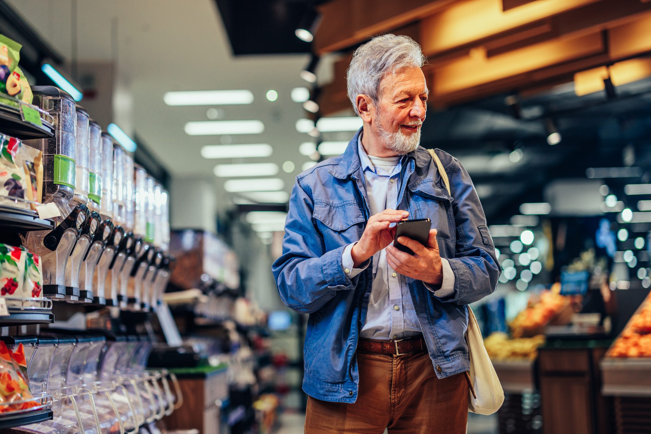 Senior man is doing grocery shopping, holding mobile phone with a shopping list and having shopping bag on his shoulder.