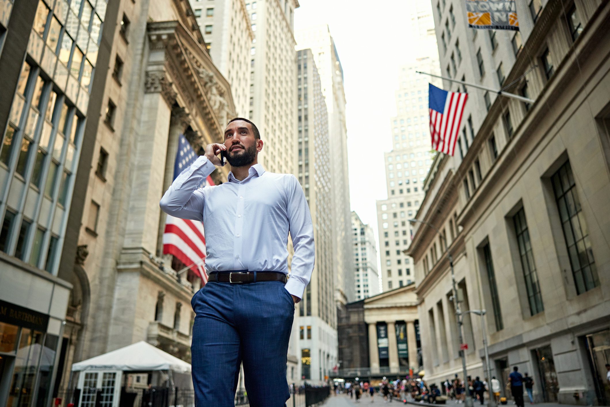 Bearded man in early 30s with hand in pocket walking in Manhattan Financial District, talking with caller, landmark financial building in background.