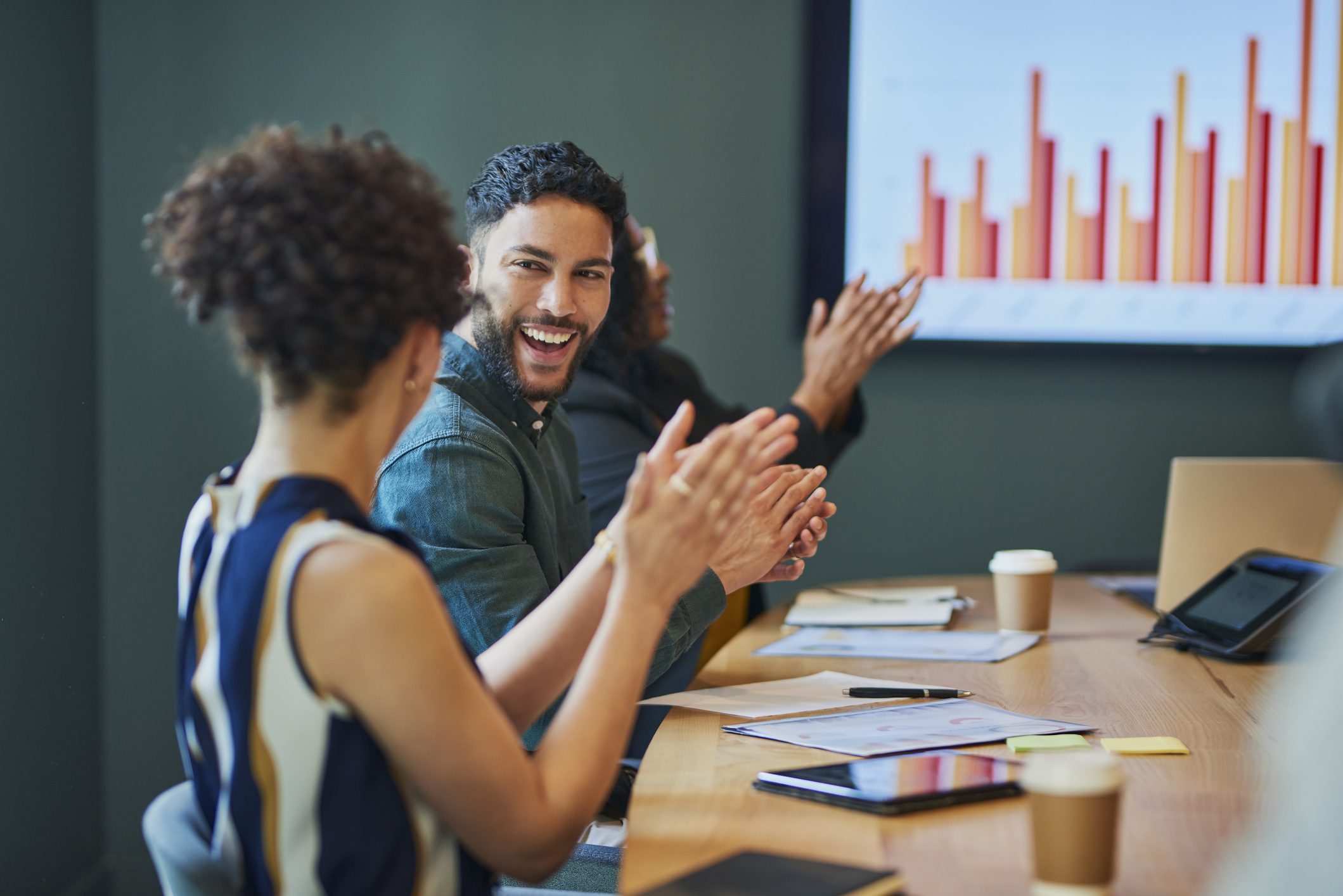 Group of business professionals clapping and smiling during a meeting in the boardroom. They are all sitting around a conference table and looking at a presentation on the screen. The people are all dressed in business casual attire and are engaged in the meeting.