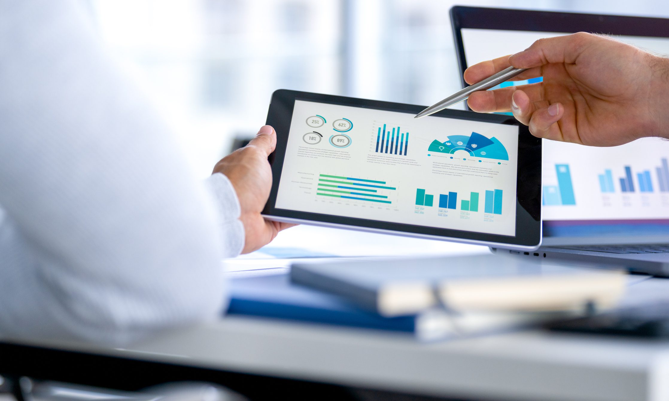 Two business men meeting and looking at financial data, charts and graphs on a digital tablet and a laptop computer. There are documents and paperwork on the table. One man is pointing at the screen