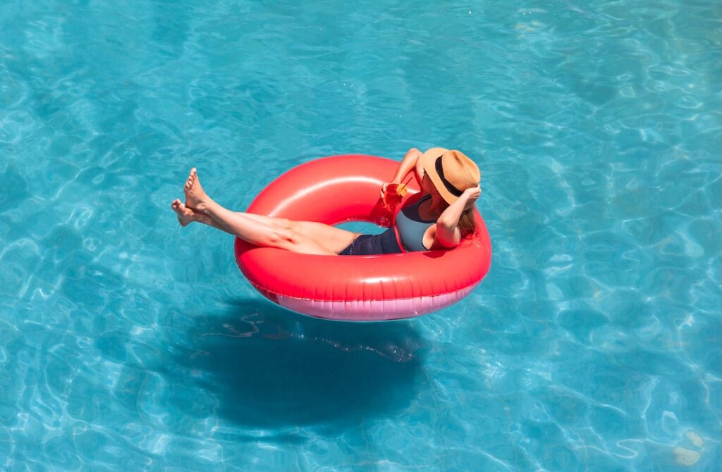 A woman enjoying a sunny day floating on a pink inflatable in a clear blue swimming pool, holding a refreshing drink.