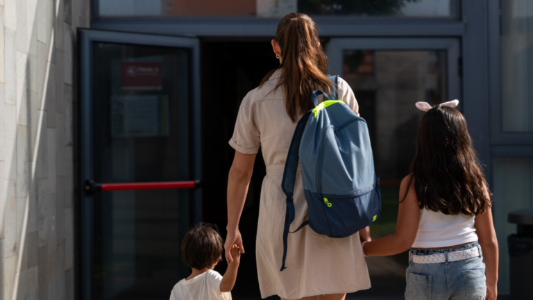 A woman is walking with two children, one of whom is wearing a backpack. The scene is casual and relaxed, with the family likely heading to school or another destination