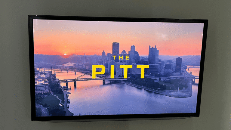 wall-mounted flat-screen TV displaying the title card of the HBO Max series The Pitt, showing an aerial view of the Pittsburgh skyline at sunset with rivers and bridges in the foreground. The show's title is displayed in bold yellow text — 'THE' in smaller letters above the large word 'PITT' — centered over the cityscape.